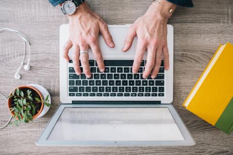 Typing on a laptop with a small potted plant, a yellow notebook and earbuds on the desk