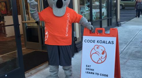 Person in Koala costume stands outside an office door with a sign that reads eat, drink, learn to code