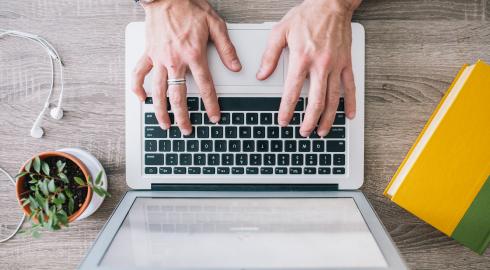 Typing on a laptop with a small potted plant, a yellow notebook and earbuds on the desk