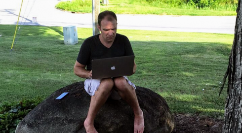 Person sitting barefoot on a rock under a tree working on a laptop